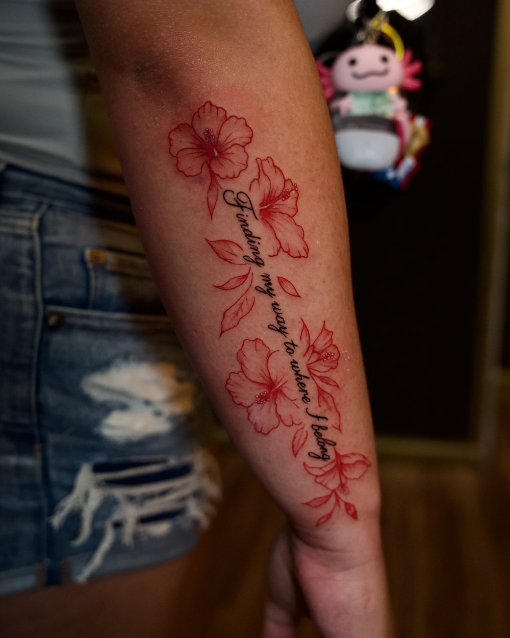 Red ink hibiscus flowers with script lettering reading Finding my way to where I belong, down the forearm, by Rodney Alacan