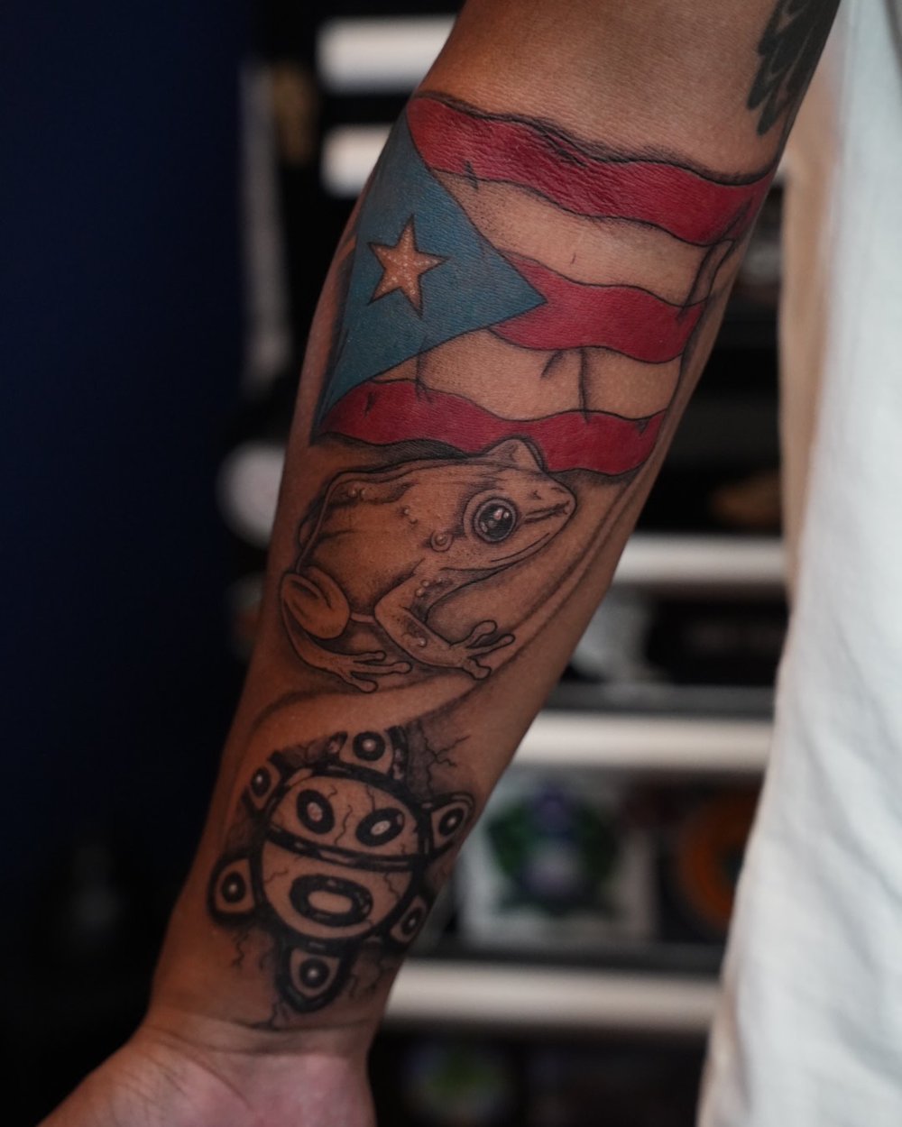 Color Puerto Rico flag with a coquí frog and taíno sun tattooed on the forearm, by Rodney Alacan